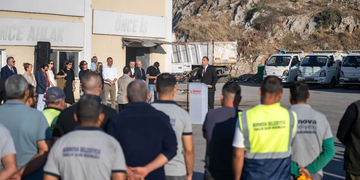 Bornova Belediyesi logolu bir kürsüde konuşma yapan takım elbiseli bir yetkili ve onu dinleyen, üzerinde Bornova Belediyesi Temizlik İşleri Müdürlüğü yazılı üniformalar giymiş çalışanlar ile belediye personeli açık havada bir araya gelmiş durumda. Arka planda 