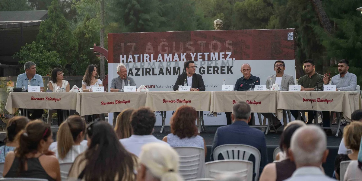 Açık havada düzenlenen bir panelde, Bornova Belediyesi logolu masaların arkasında oturan dokuz konuşmacı, kendilerini dinleyen bir izleyici grubuna hitap ediyor. Konuşmacıların arkasındaki büyük panoda 