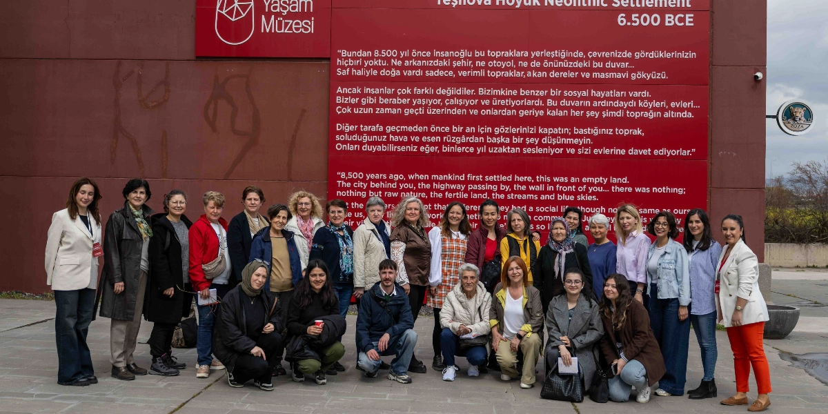 Bornova Yeşilova Höyüğü Ziyaretçi Merkezi'nin kırmızı renkli dış duvarı önünde poz veren, çoğunluğu kadınlardan oluşan kalabalık bir grup. Duvarın üzerinde 