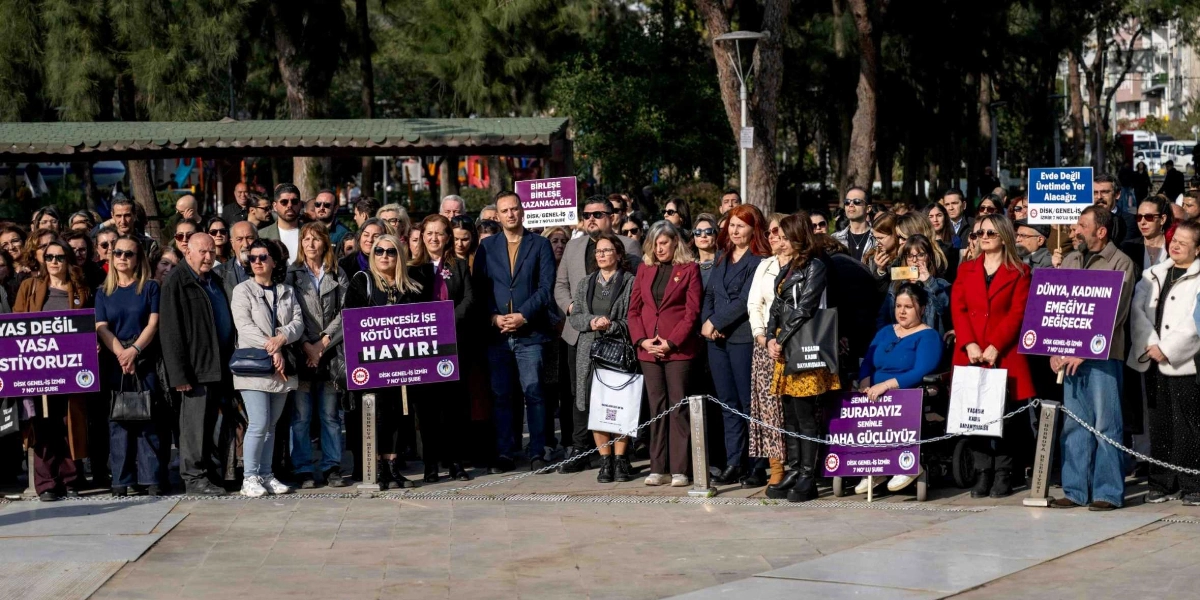 Açık havada, park benzeri bir alanda bir araya gelmiş, ellerinde DİSK Genel-İş İzmir 7 No'lu Şube imzalı pankartlar taşıyan kalabalık bir grup. Pankartlarda 