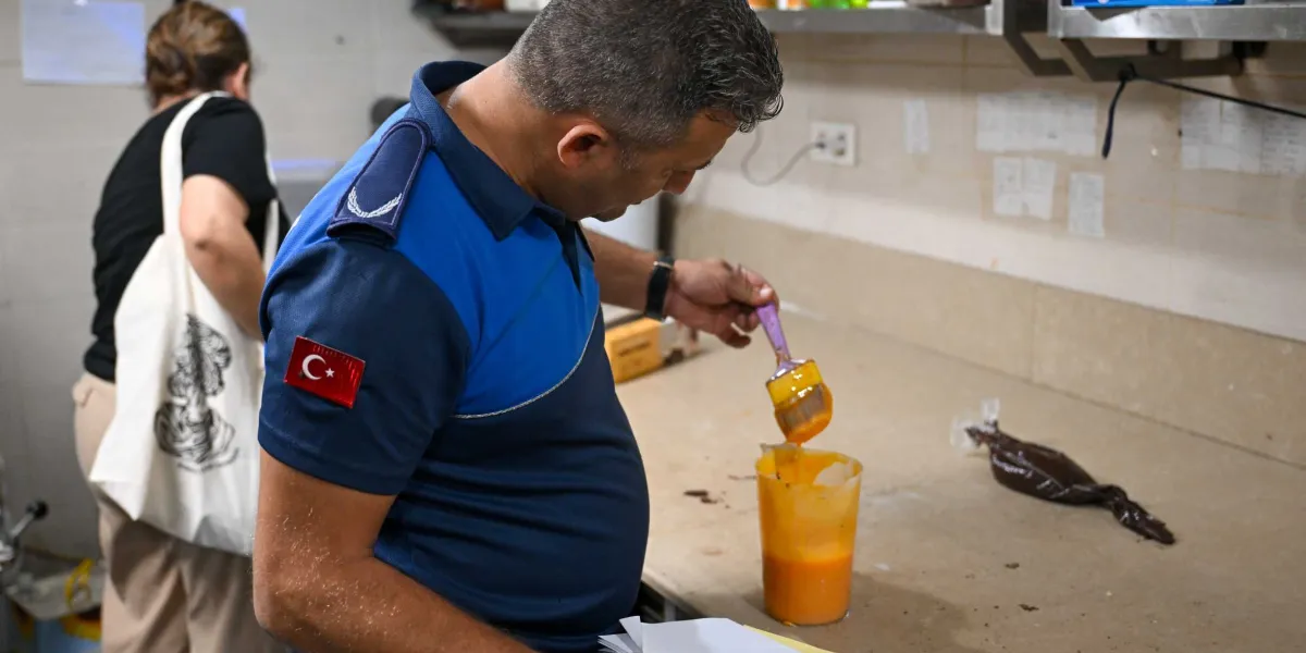 Mavi üniformalı bir zabıta memuru, bir fırın tezgahı üzerinde bulunan turuncu renkli bir karışımı fırça yardımıyla inceliyor. Memurun kolunda Türk bayrağı arması bulunuyor ve arka planda beyaz tişörtlü bir kadın ayakta duruyor.