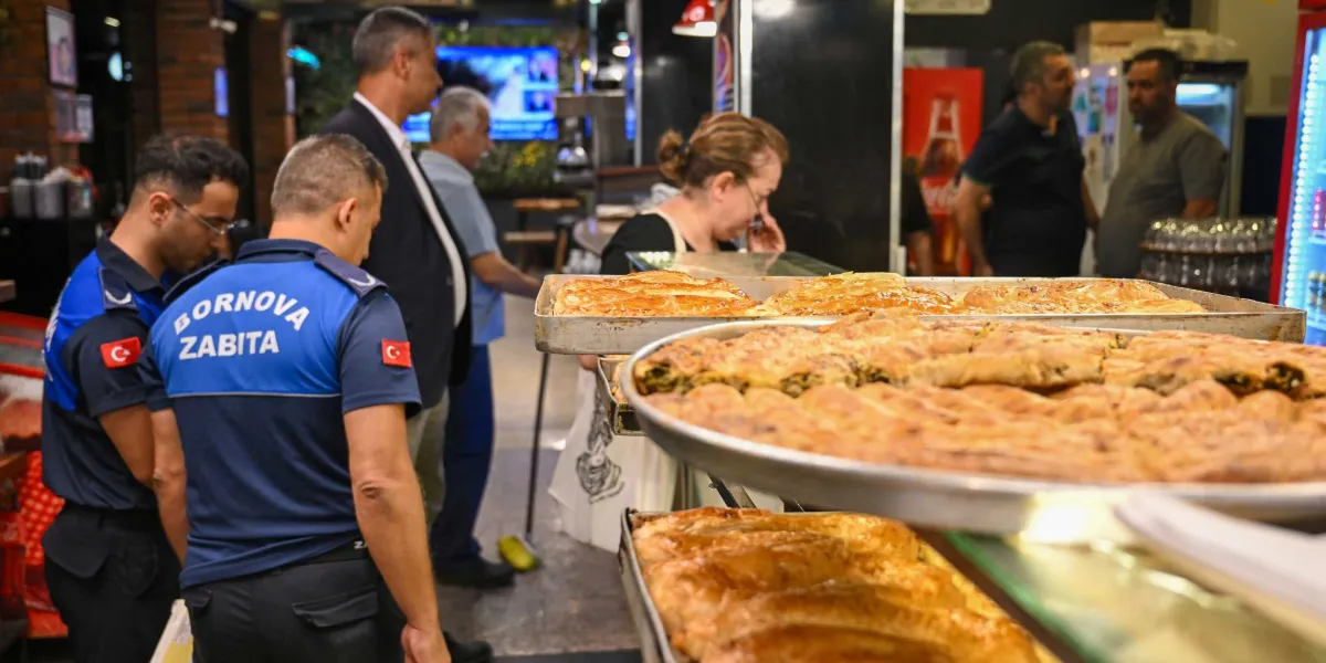 Bornova Belediyesi zabıta ekipleri, bir fırında denetim yapıyor. Ön planda metal tepsiler içinde taze pişmiş börekler yer alırken, arka planda zabıta üniformalı görevliler ve işletme çalışanları bulunuyor.