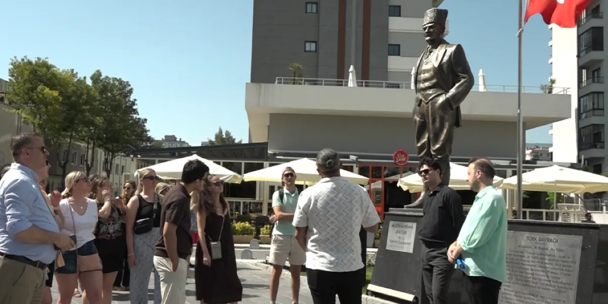 Bornova'da bir meydanda, Mustafa Kemal Atatürk'ün bronz heykelinin önünde toplanmış, farklı yaş ve giyim tarzlarına sahip bir grup genç ve yetişkin, rehberlerini dinliyor. Heykelin kaidesinde 