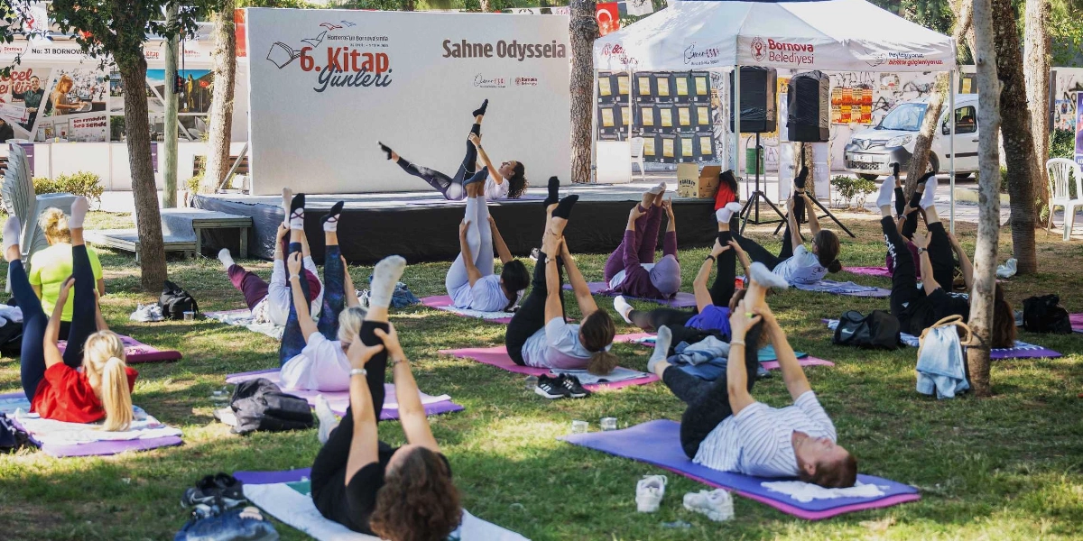 Bornova Belediyesi 6. Kitap Günleri kapsamında, ağaçlık bir alanda yoga matları üzerinde egzersiz yapan bir grup kadın. Arka planda 