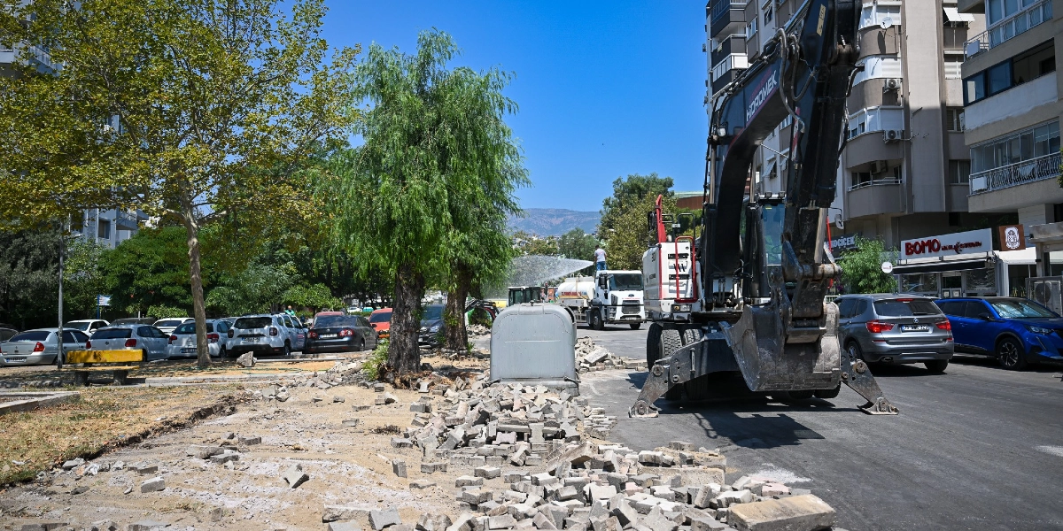 Bornova'da bir cadde üzerinde devam eden yol ve kaldırım yenileme çalışması. Sağ tarafta büyük bir Hidromek iş makinesi park halindeyken, arka planda bir su tankeri ağaçları suluyor. Yol kenarında sökülmüş parke taşları, park halindeki araçlar ve çevredeki apartmanlar görülüyor.