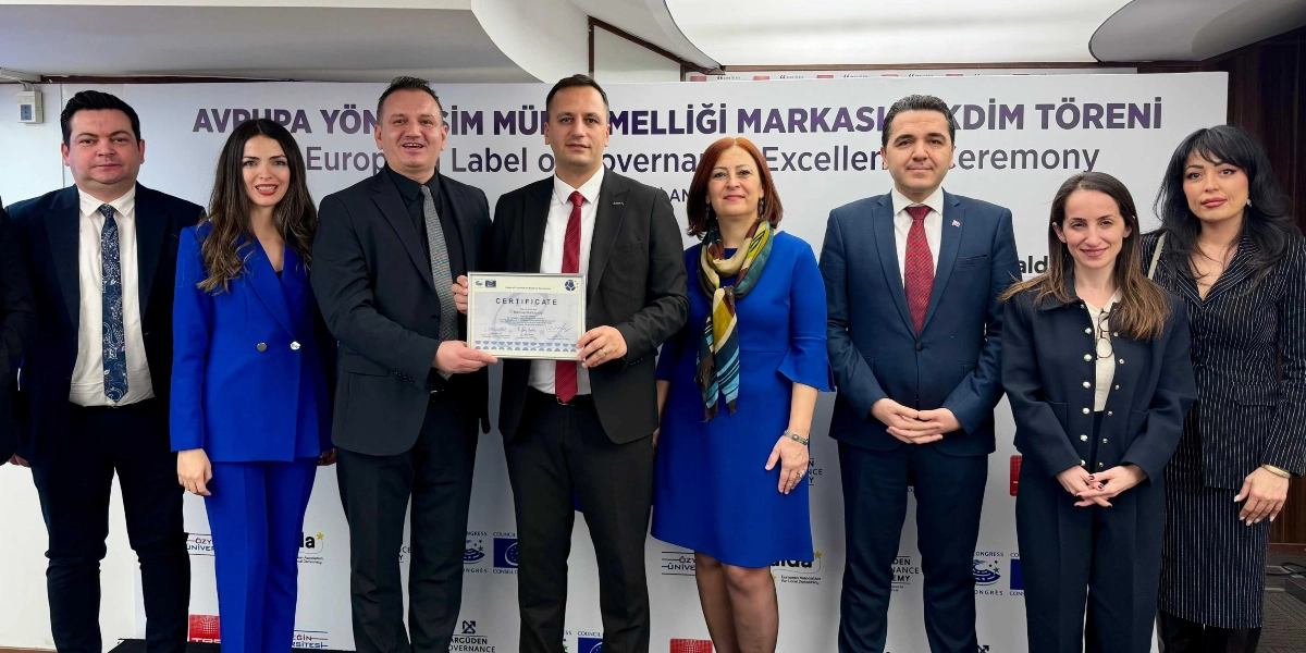 Avrupa Yönetişim Mükemmelliği Markası Takdim Töreni'nde, ellerinde sertifika tutan bir yetkilinin de bulunduğu sekiz kişilik grup, arkalarında törenin adının yazılı olduğu bir fon önünde yan yana durarak poz veriyor. Gruptaki erkekler takım elbise ve kravat, kadınlar ise şık ceket ve elbiseler giyiyor. Arka planda Avrupa Konseyi ve çeşitli kurumların logoları yer alıyor.