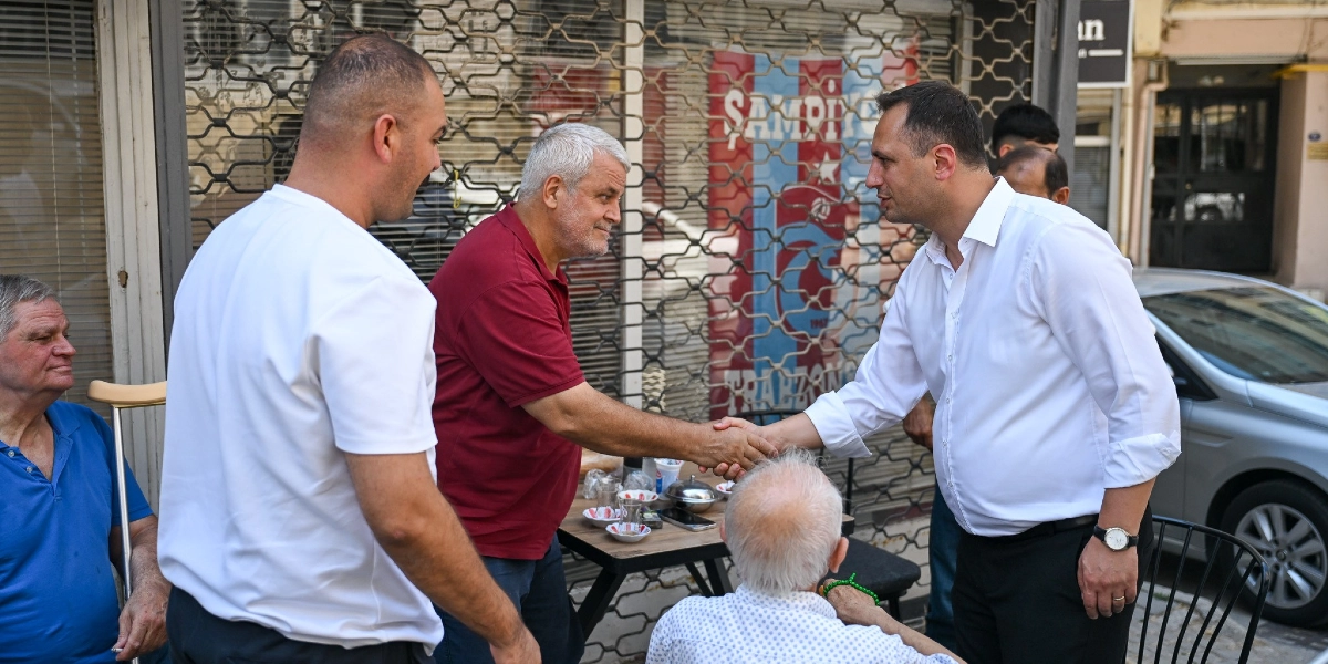 Beyaz gömlekli bir erkek, bordo polo yaka tişört giyen başka bir erkekle el sıkışıyor. Arka planda kepenkleri kapalı bir dükkan ve önünde masada oturan diğer kişiler bulunuyor. Masada çay bardakları ve küçük bir tabak yer alıyor. Sol tarafta koltuk değneğiyle oturan mavi tişörtlü bir kişi, sağ tarafta ise sırtı dönük oturan beyaz saçlı bir kişi görülüyor.