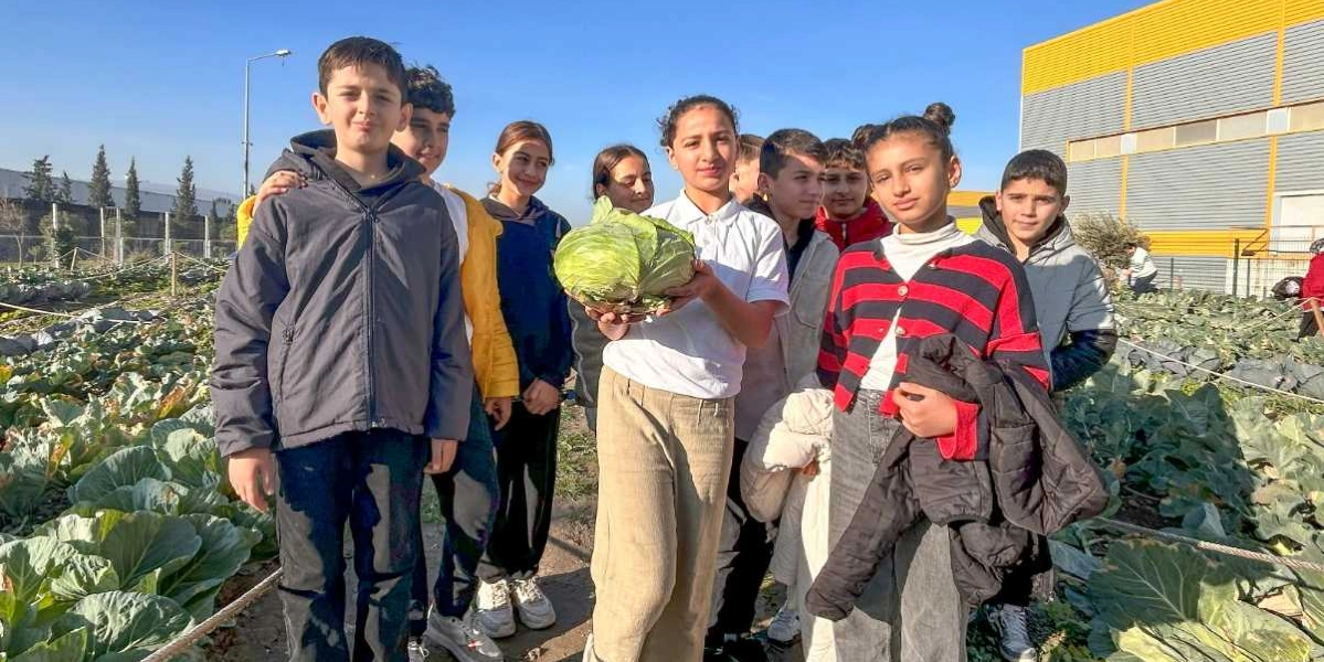 Açık havada, lahana tarlasında yan yana duran bir grup öğrenci, ellerinde tuttukları büyük bir lahanayı gösteriyor. Öğrenciler günlük kıyafetleriyle, güneşli bir günde tarlanın ortasında objektife gülümsüyor. Arka planda ise modern bir bina yapısı ve ağaçlar yer alıyor.