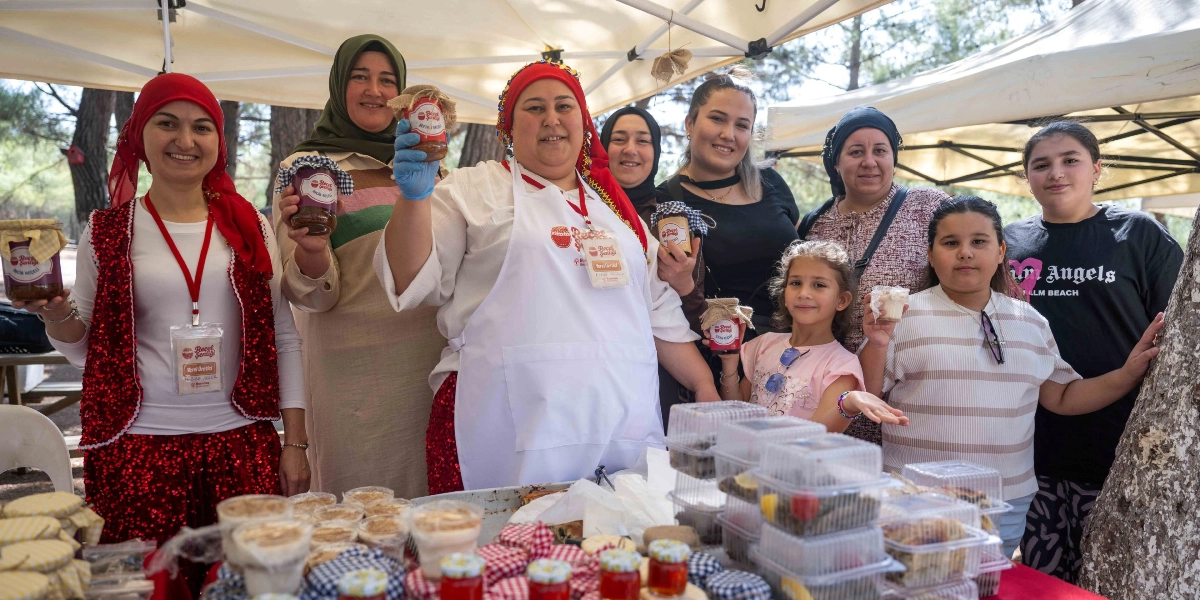 Açık havada, beyaz tentelerin altında kurulan stantta, kendi yaptıkları reçelleri sergileyen ve gülümseyerek kameraya poz veren bir grup kadın ve çocuk. Kadınlardan bazıları geleneksel kıyafetler ve başörtüsü takarken, önlerinde reçel kavanozları ve plastik kaplarda paketlenmiş yiyecekler bulunuyor. Ortadaki kadın beyaz bir önlük giyiyor ve elinde 