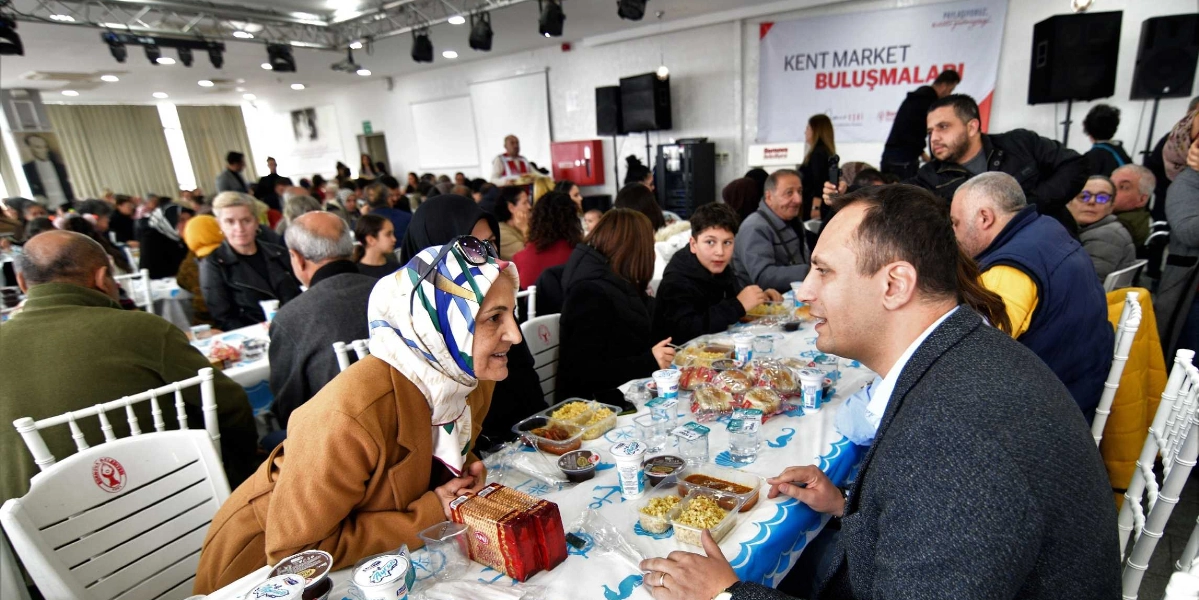 Bornova Belediye Başkanı Ömer Eşki, kalabalık bir salonda düzenlenen yemekte, masada oturan başörtülü bir kadınla sohbet ediyor. Masanın üzerinde plastik kaplarda yemekler, su ve paketli gıdalar bulunuyor. Arka planda 