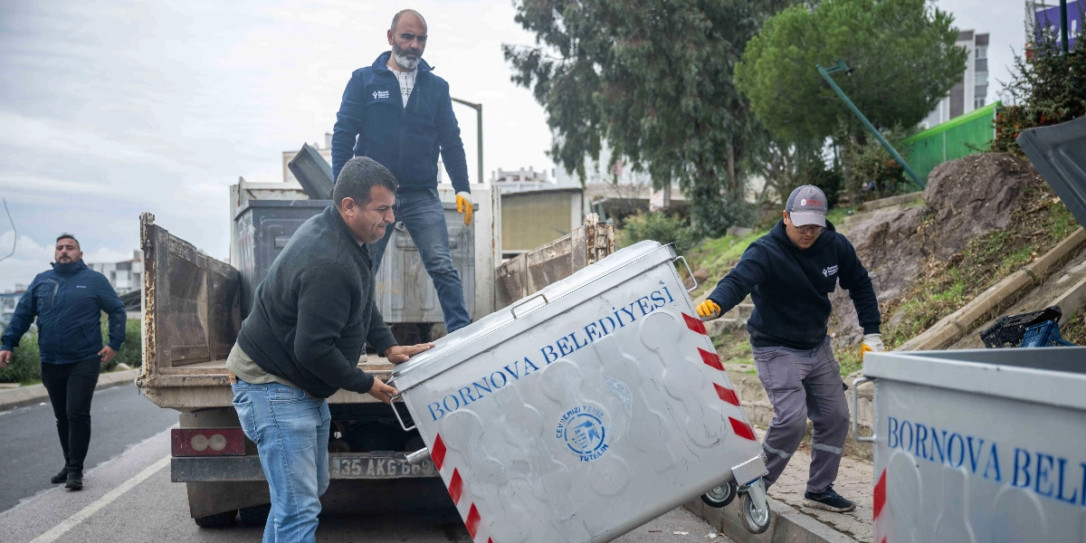 Bornova Belediyesi personeli, üzerinde belediyenin logosu ve ismi bulunan gri renkli yeni çöp konteynerlerini bir kamyondan indirerek kaldırıma yerleştiriyor.