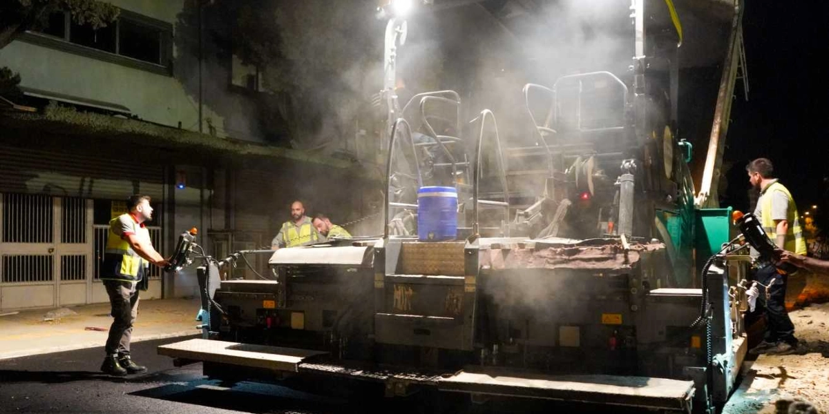 Gece saatlerinde bir sokakta asfalt serme makinesiyle yapılan yol yenileme çalışması. İş makinelerinin etrafında sarı reflektörlü yelek giymiş işçiler çalışıyor ve makineden yoğun bir duman yükseliyor.