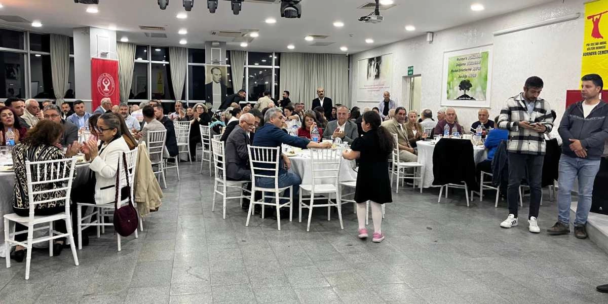 Aydınlık bir salonda düzenlenen yemekli toplantıda, beyaz örtülü yuvarlak masaların etrafında oturan çok sayıda katılımcı yer alıyor. Salonun duvarlarında Bornova Belediyesi'nin logosu, Mustafa Kemal Atatürk'ün portresi ve Pir Sultan Abdal Kültür Derneği Bornova Cemevi'ne ait sarı renkli bir flama bulunuyor. Katılımcıların bir kısmı masalarda sohbet ederken, siyah elbiseli küçük bir kız çocuğu salonun ortasında ayakta duruyor. Sağ tarafta ise ayakta duran iki erkek katılımcı, topluluğa doğru bakıyor.