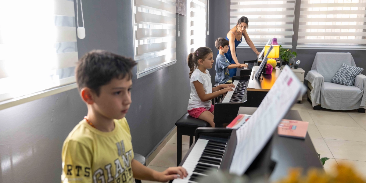 Aydınlık bir sınıfta piyano eğitimi alan üç çocuk ve onlara rehberlik eden bir eğitmen. Ön planda sarı tişörtlü bir erkek çocuk piyano çalarken, arka planda bir kız çocuğu ve başka bir erkek çocuk daha piyano başında oturuyor. Mavi elbiseli kadın eğitmen, çocuklara eşlik ederek piyano dersini yönetiyor. Sınıfta piyanoların yanı sıra, pencerelerde stor perdeler ve köşede gri bir koltuk bulunuyor.