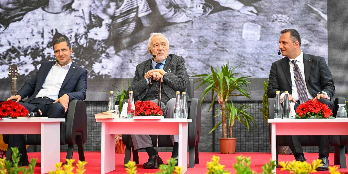 Üç erkek, sahnede yan yana yerleştirilmiş koltuklarda oturuyor. Ortada, elinde bastonuyla oturan tarihçi İlber Ortaylı, sağında ve solunda ise takım elbiseli iki yetkili bulunuyor. Her birinin önünde beyaz masalar, masaların üzerinde ise kırmızı çiçek aranjmanları ve su şişeleri yer alıyor. Arka planda, üzerinde siyah beyaz bir fotoğrafın olduğu büyük bir ekran bulunuyor.