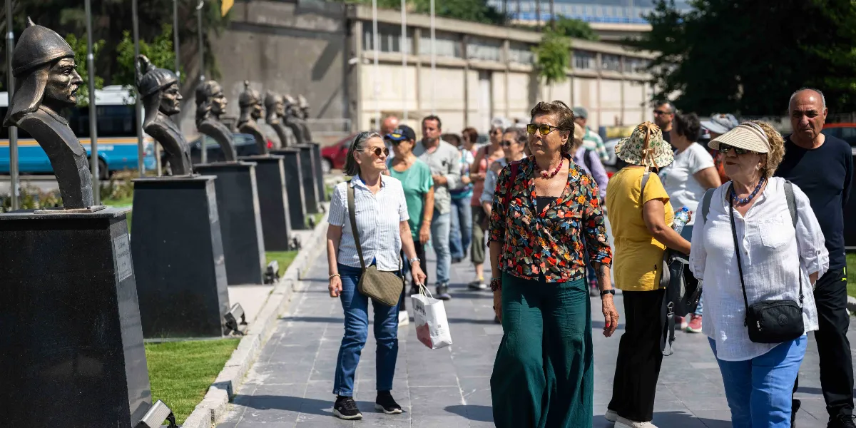 Bornova'da bir park yolunda yürüyen bir grup insan, yanlarında sıralanmış tarihi figürleri temsil eden büstlerin önünden geçiyor. Büstler, kaideler üzerinde arka arkaya dizilmiş şekilde yer alıyor. İnsanlar gündelik kıyafetler içinde, güneşli bir havada yürüyüş yaparken görülüyor.