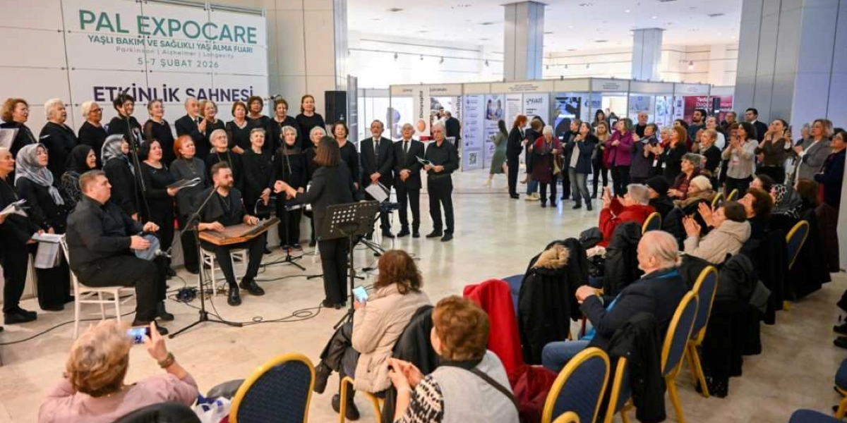 Pal Expocare Yaşlı Bakım ve Sağlıklı Yaş Alma Fuarı'nın etkinlik sahnesinde, bir koro ve kanun çalan bir müzisyen performans sergiliyor. Siyah kıyafetli kadınlardan oluşan koro, bir şef yönetiminde şarkı söylerken, izleyiciler fuar alanındaki sandalyelerde oturarak onları dinliyor. Arka planda fuar stantları ve katılımcılar yer alıyor.