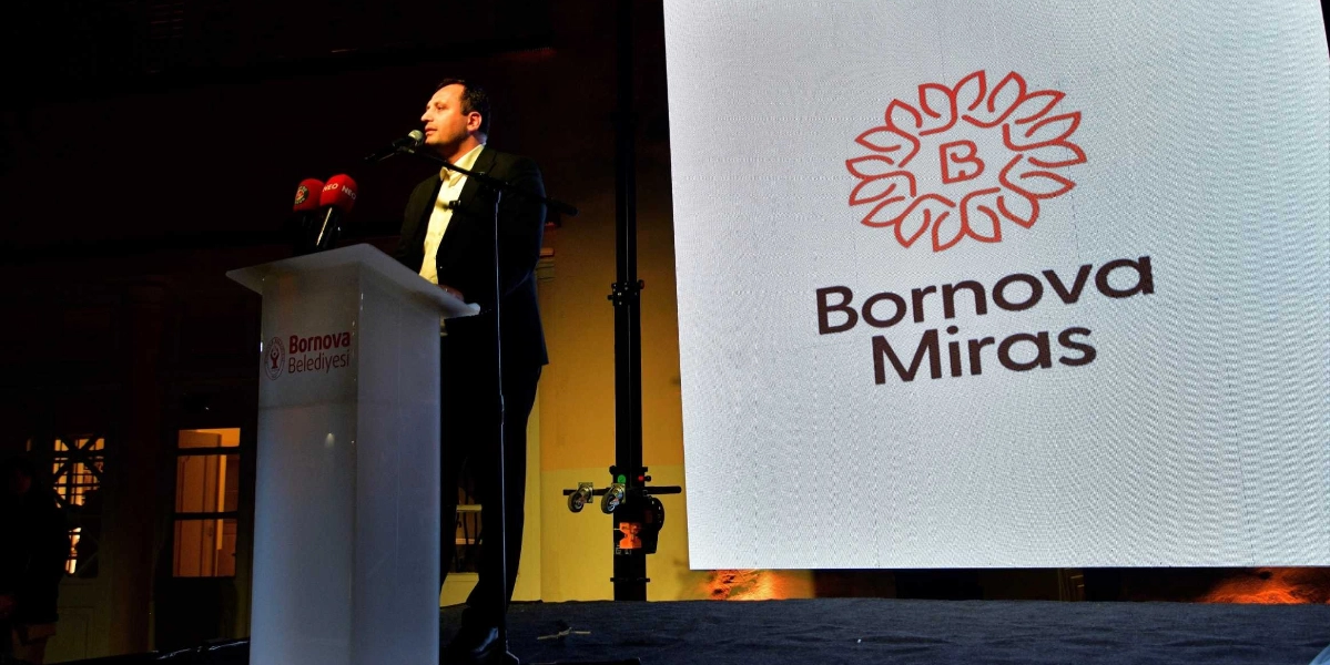 Koyu renkli takım elbiseli bir erkek, üzerinde Bornova Belediyesi logosu bulunan şeffaf bir kürsüde mikrofon başında konuşma yapıyor. Konuşmacının arkasında yer alan büyük ekranda, stilize edilmiş bir çiçek figürü ve altında 