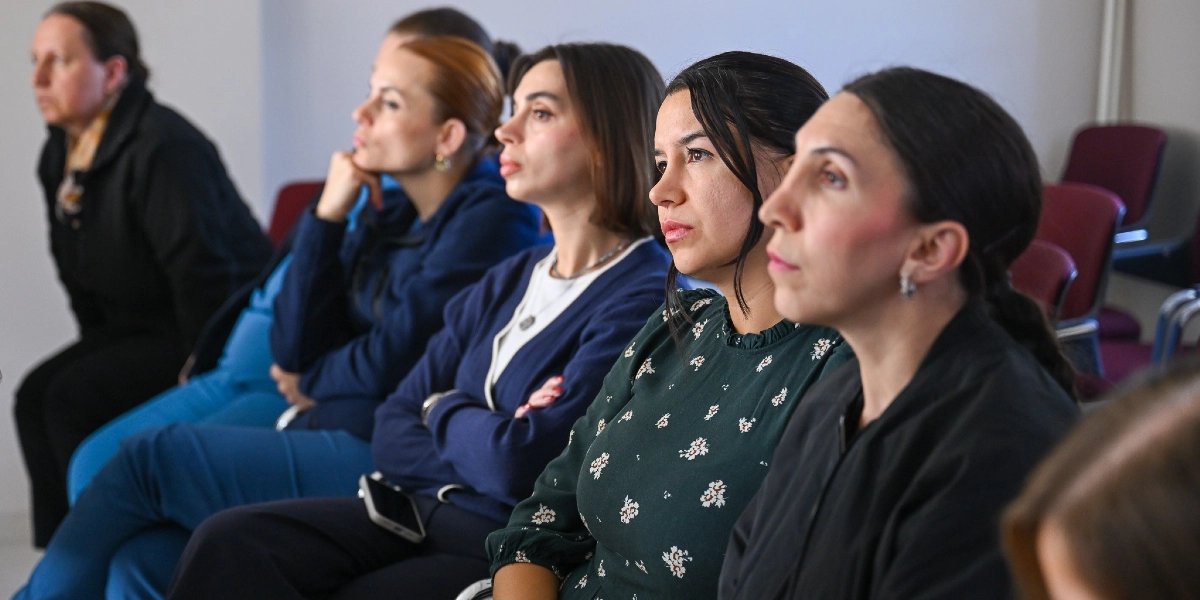 Bir konferans salonunda yan yana dizilmiş sandalyelerde oturan ve dikkatle karşıya bakan bir grup kadın. Kadınlar farklı renklerde günlük kıyafetler giymiş durumdalar; aralarında koyu yeşil çiçek desenli bir elbise giyen, lacivert hırkalı ve siyah kıyafetli kişiler bulunuyor. Katılımcılar ciddi ve odaklanmış bir ifadeyle bir sunumu veya konuşmacıyı dinliyorlar.