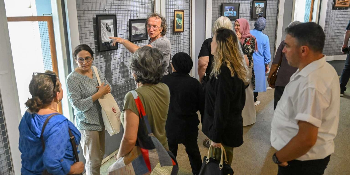 Bir sanat galerisinde, duvardaki metal ızgaralara asılı fotoğrafları inceleyen bir grup insan. Kıvırcık saçlı orta yaşlı bir adam, duvardaki siyah beyaz bir fotoğrafı eliyle işaret ederek yanındaki kadına bir şeyler anlatıyor. Çevredeki diğer katılımcılar ise farklı eserleri dikkatle inceliyor; aralarında başörtülü kadınların da bulunduğu grup, sanat eserleri üzerine sohbet ederek galeriyi geziyor.