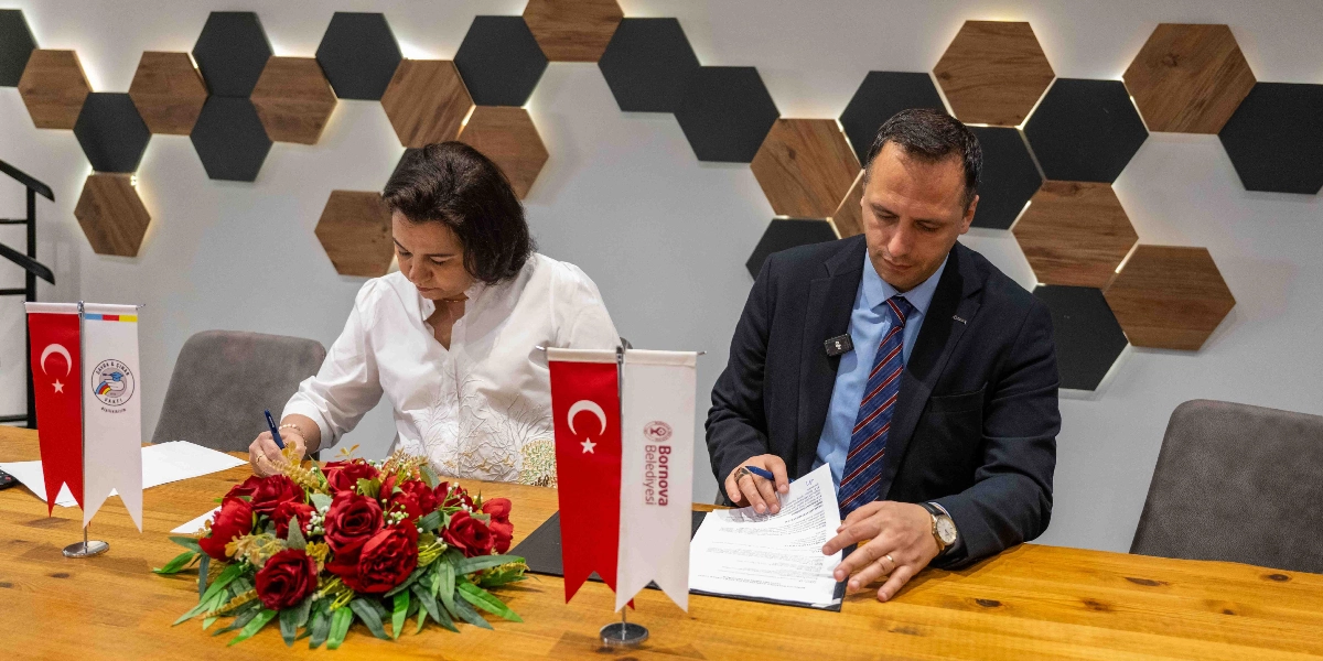 Bornova Belediye Başkanı ve Sayra & Çınar Vakfı temsilcisi, ahşap bir masa başında oturarak iş birliği protokolünü imzalıyor. Masanın üzerinde Türk bayrakları, kurum logoları ve kırmızı güllerden oluşan bir aranjman bulunuyor. Arka planda ise altıgen ahşap panellerle dekore edilmiş modern bir duvar yer alıyor.
