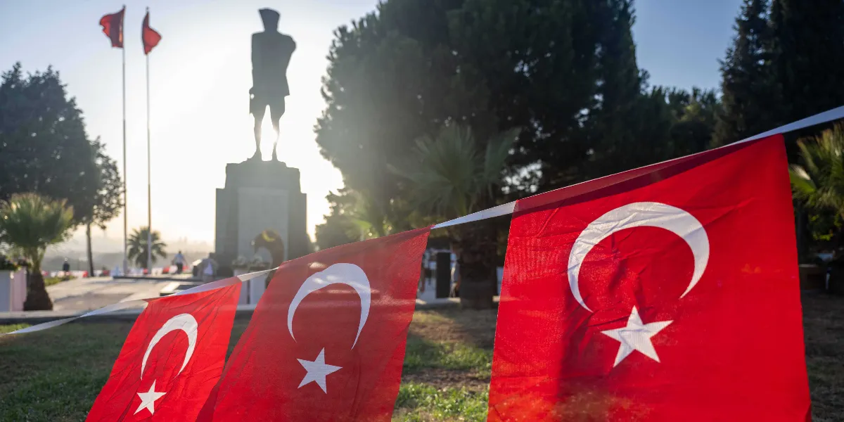 Arka planda güneş ışığıyla silüeti belirginleşen Atatürk heykeli ve yanındaki direklerde dalgalanan Türk bayrakları yer alıyor. Ön planda ise bir ipe dizilmiş, yan yana sıralanan Türk bayrakları odak noktasında bulunuyor. Çevrede ağaçlar ve park alanı görülüyor.
