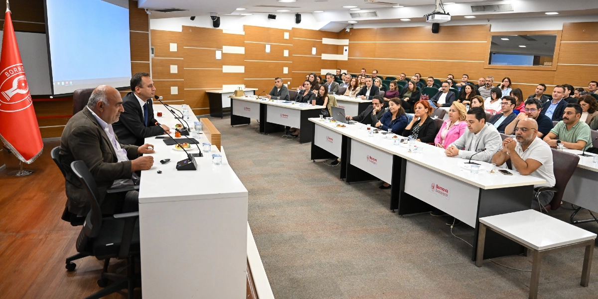 Bornova Belediyesi'ne ait bir toplantı salonunda, U şeklinde düzenlenmiş masalarda oturan kalabalık bir grup katılımcı ve masanın başında konuşma yapan iki yetkili. Salonun sol tarafında Bornova Belediyesi'nin kırmızı bayrağı, arka planda ise sunum için kullanılan büyük bir ekran yer alıyor. Katılımcılar dikkatle dinlerken, masaların üzerinde Bornova Belediyesi logoları ve içecekler bulunuyor.
