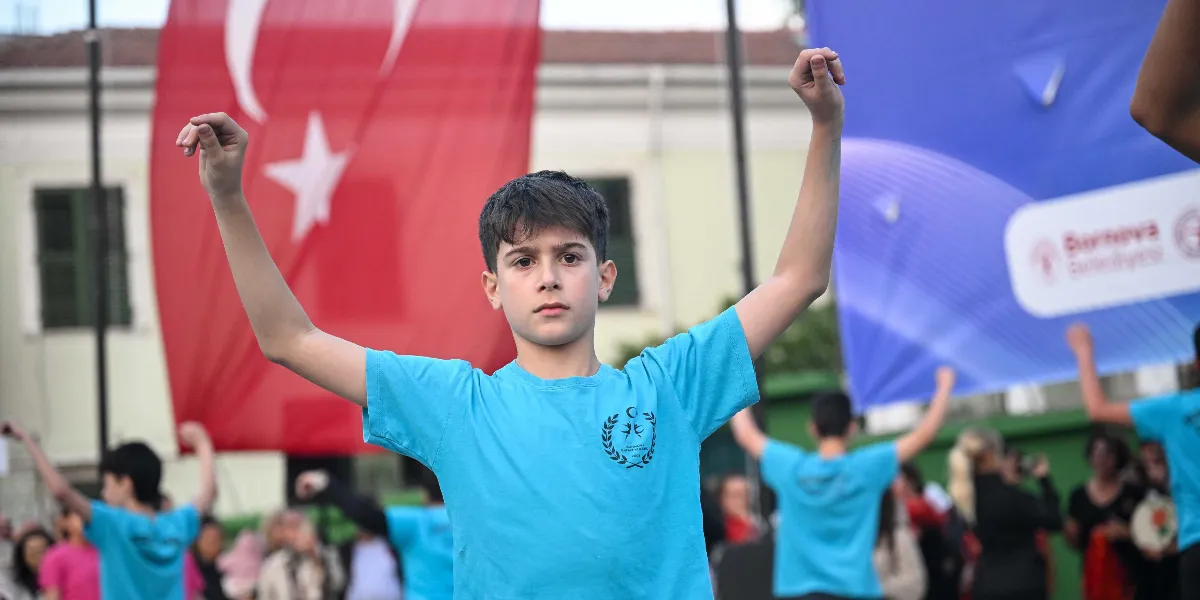 Açık havada düzenlenen Dünya Dans Günü etkinliğinde, üzerinde Bornova Belediyesi logosu bulunan açık mavi tişörtlü genç bir erkek çocuğu, kolları havada dans figürü sergiliyor. Arka planda Türk bayrağı ve üzerinde Bornova Belediyesi logosu bulunan mavi bir pankart yer alırken, çevresinde benzer kıyafetlerle dans eden diğer çocuklar ve izleyiciler görülüyor.
