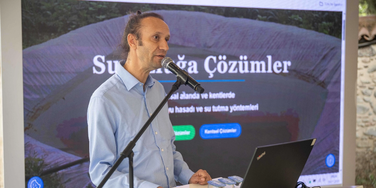 Açık mavi gömlekli, saçları arkadan toplanmış bir erkek, önünde mikrofon bulunan bir kürsüde konuşma yapıyor. Konuşmacının arkasında, üzerinde 
