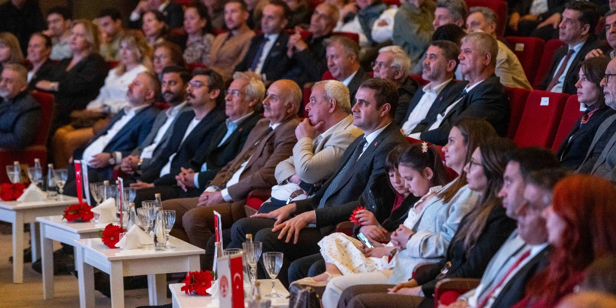 Konferans salonunda kırmızı koltuklarda oturan, resmi kıyafetli kalabalık bir izleyici grubu sahneye doğru bakıyor. Ön sıralardaki beyaz masaların üzerinde küçük Türk bayrakları, bardaklar ve çiçek süslemeleri bulunuyor. İzleyiciler arasında Bornova Belediye Başkanı Mustafa İduğ'un da yer aldığı, dikkatle dinleyen ve izleyen bir topluluk görülüyor.