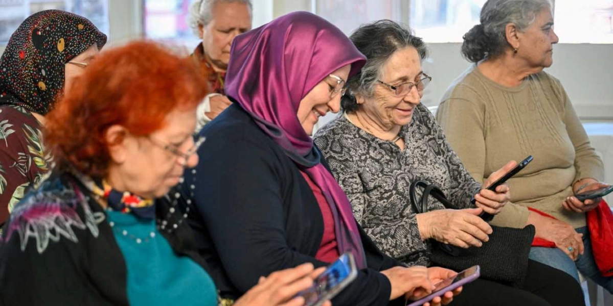 Bir sınıfta yan yana oturan, farklı yaş gruplarından kadınlar ellerindeki akıllı telefonları inceliyor. Ön planda mor başörtülü, gülümseyen bir kadın ve yanında turuncu saçlı bir kadın telefon ekranlarına odaklanmış durumda. Arka planda ise gözlüklü ve gri saçlı kadınlar ile başörtülü diğer katılımcılar dikkatle telefonlarını kullanıyor.