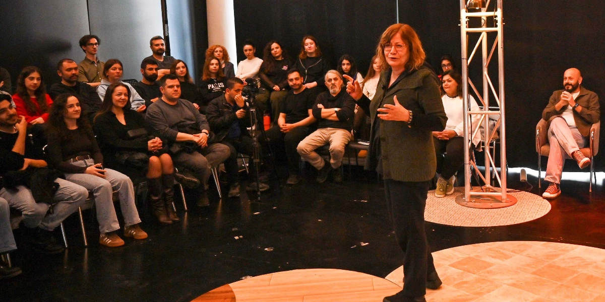 Koyu renkli bir sahnede, elinde kalem tutan orta yaşlı bir kadın ayakta konuşma yaparken, karşısında yarım daire şeklinde oturmuş yaklaşık yirmi kişilik bir izleyici grubu onu dikkatle dinliyor. Sahnenin sağ tarafında metal bir ışık kulesi ve oturmuş vaziyette bir erkek katılımcı bulunuyor. İzleyiciler arasında farklı yaş gruplarından kadın ve erkekler yer alıyor; ortamda profesyonel bir sunum veya söyleşi havası hakim.
