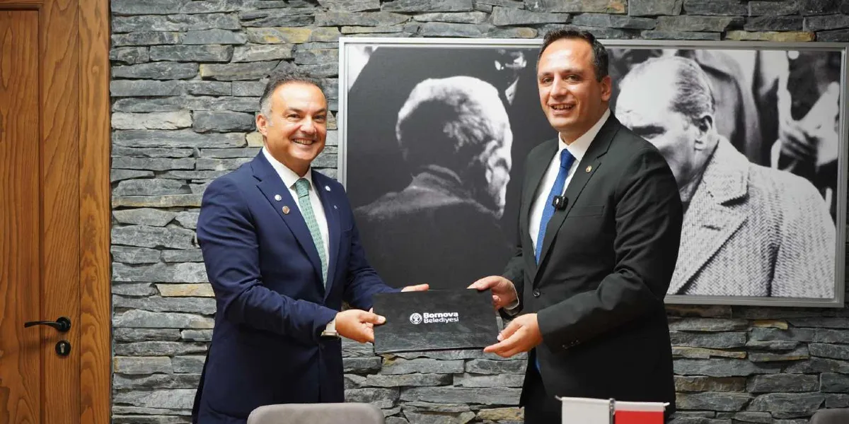 Takım elbiseli iki erkek, üzerinde Bornova Belediyesi logosu bulunan siyah bir dosyayı birlikte tutarak gülümseyerek poz veriyor. Arka planda taş duvar üzerinde Mustafa Kemal Atatürk'ün siyah beyaz bir portresi yer alıyor.