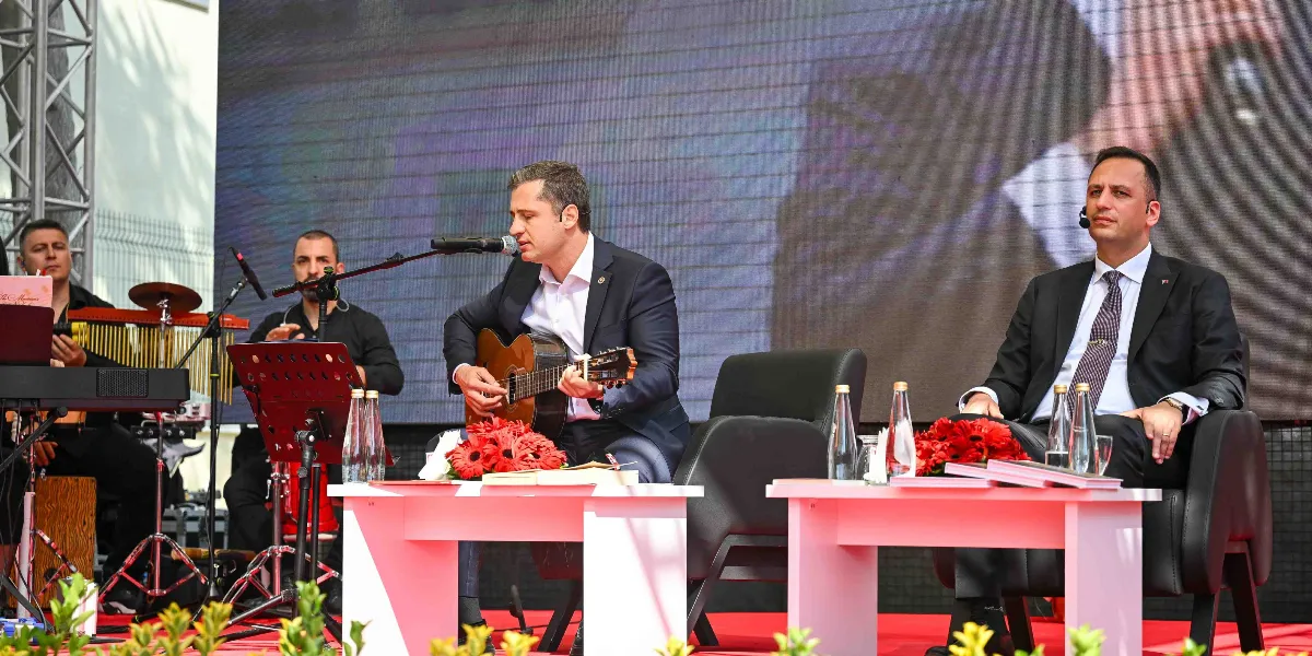 Açık havada kurulan bir sahnede, takım elbiseli bir kişi gitar çalıp şarkı söylerken, yanında başka bir takım elbiseli kişi oturarak onu dinliyor. Sahnenin sol tarafında müzik aletleri çalan iki kişi daha bulunuyor. Önlerinde beyaz masalar ve kırmızı çiçekler yer alıyor, arka planda ise büyük bir ekran bulunuyor.