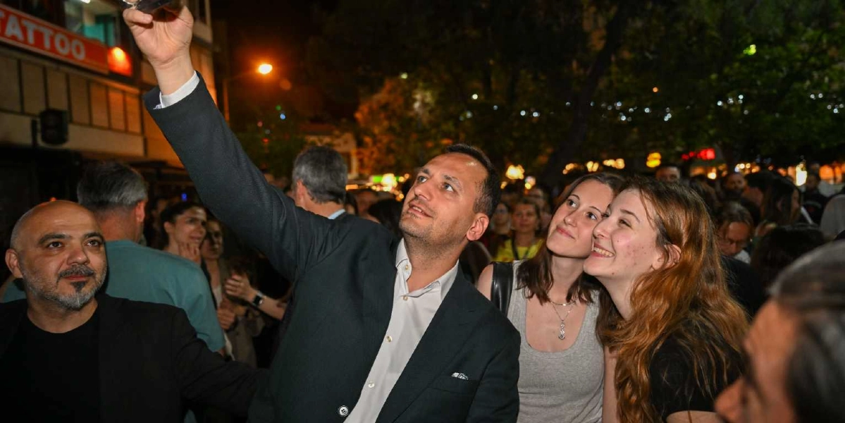 Gece vakti kalabalık bir sokakta, koyu renk ceketli bir adamın etrafındaki iki genç kadınla birlikte gülümseyerek selfie çektiği an.