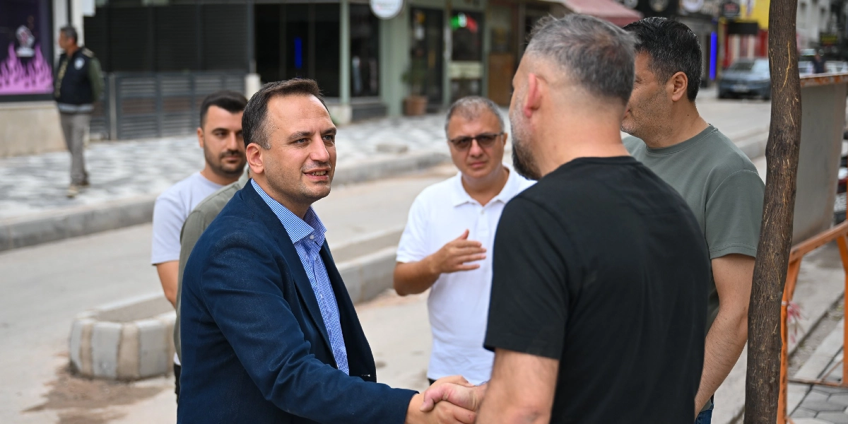 Bornova Zafer Caddesi'nde lacivert ceketli ve mavi gömlekli bir yetkili, siyah tişörtlü bir vatandaşla tokalaşırken çevresindeki diğer kişilerle sohbet ediyor. Arka planda düzenleme çalışmaları devam eden cadde ve dükkanlar görülüyor.