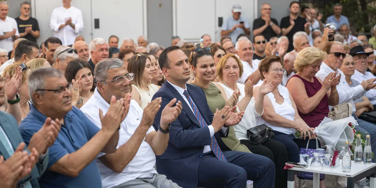 Açık havada düzenlenen bir etkinlikte, yan yana oturmuş ve alkışlayan bir grup insan. Ön planda, lacivert takım elbiseli ve çizgili kravatlı bir erkek ile yanında oturan yeşil bluzlu bir kadın dikkat çekiyor. Hemen yanlarında beyaz gömlekli ve gözlüklü bir erkek ile mavi polo yaka tişörtlü bir başka erkek de alkışlayarak etkinliğe katılıyor. Arka planda ise farklı yaş gruplarından çok sayıda katılımcı, benzer şekilde alkışlayarak izleyici koltuklarında oturuyor. Önlerinde üzerinde su şişeleri ve bardaklar bulunan küçük bir masa yer alıyor.