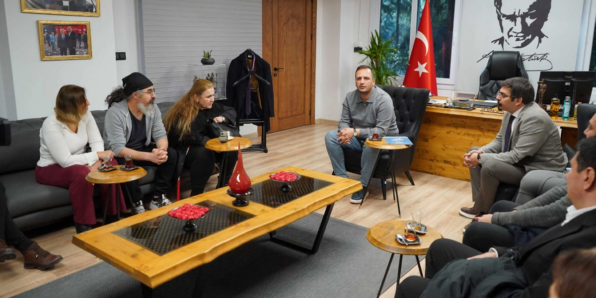 Belediye başkanı ve beraberindeki heyet, bir ofis ortamında karşılıklı oturarak toplantı yapıyor. Odanın merkezinde ahşap bir orta sehpa, etrafında ise gri bir koltuk takımı bulunuyor. Arka planda duvarda Atatürk portresi ve Türk bayrağı yer alıyor. Katılımcılar gündelik ve resmi kıyafetlerle oturmuş, sohbet ediyorlar; masaların üzerinde çay bardakları bulunuyor.