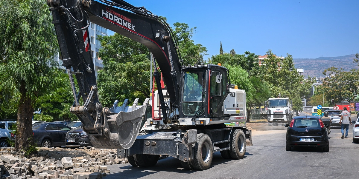 Bornova Belediyesi'ne ait Hidromek marka siyah bir iş makinesi, yol kenarındaki kaldırım düzenleme çalışması sırasında caddede park halindeki araçların yanında duruyor. Arka planda ağaçlar, diğer araçlar ve şehir dokusu görülüyor.