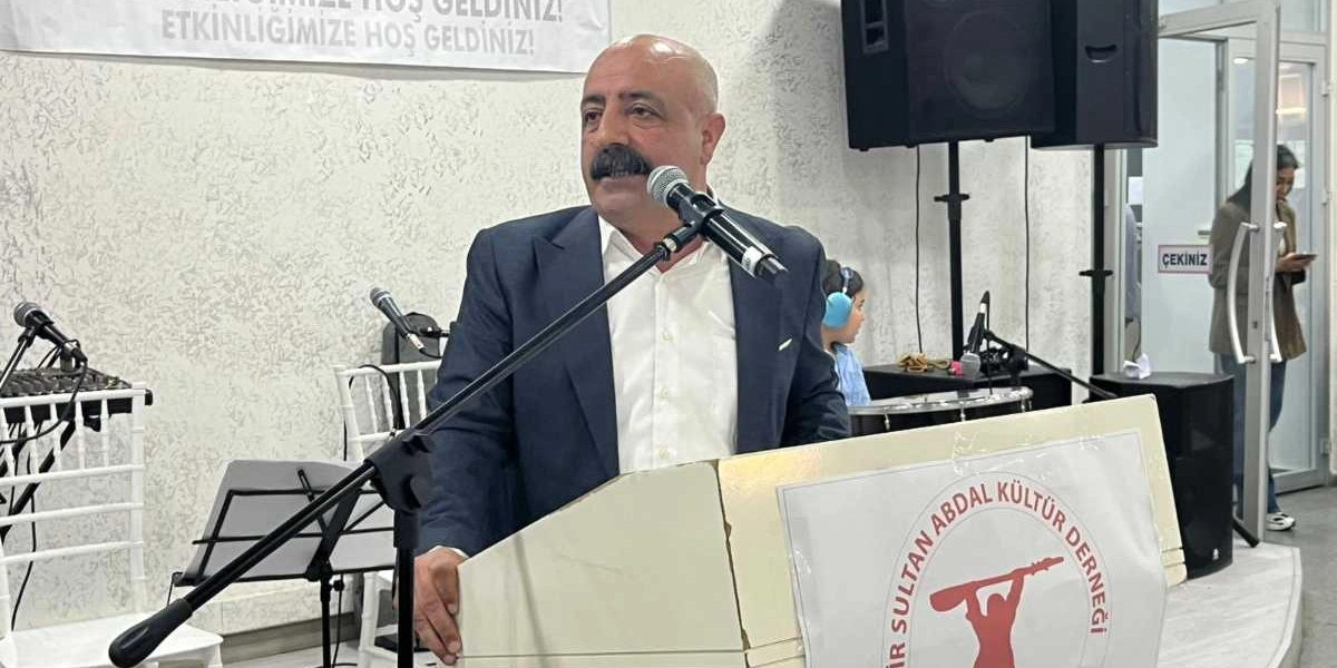 Koyu renk ceket ve beyaz gömlek giymiş, bıyıklı orta yaşlı bir erkek, üzerinde Pir Sultan Abdal Kültür Derneği logosu bulunan bir kürsüde mikrofon başında konuşma yapıyor. Arka planda beyaz bir duvar, ses sistemi ekipmanları ve etkinlik alanının girişinde duran bir kadın görülüyor.