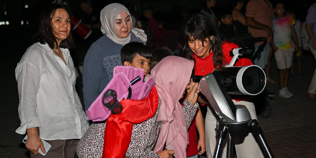 Gece vakti dışarıda kurulan bir teleskoba bakan insanlar. Kırmızı tişörtlü bir kadın teleskobun ayarlarını yaparken, pembe başörtülü bir kişi teleskobun merceğine bakıyor. Yanlarında onları izleyen, beyaz gömlekli ve gri başörtülü iki kadın ile küçük bir erkek çocuğu bulunuyor. Arka planda ise bulanık bir şekilde ayakta duran diğer insanlar yer alıyor.