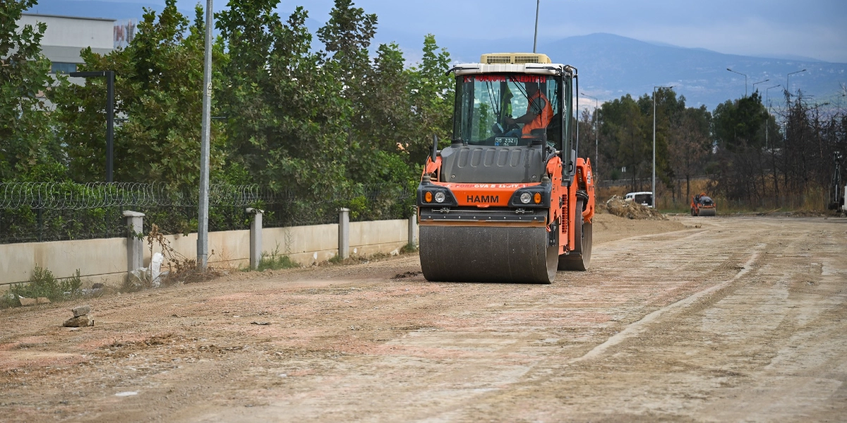 Bornova Belediyesi'ne ait turuncu renkli bir silindir, toprak ve stabilize zemin üzerinde yol çalışması yapıyor. Operatör kabininde bir çalışan bulunan iş makinesi, düz bir hat üzerinde ilerleyerek zemini sıkıştırıyor. Arka planda ağaçlar, çevre duvarı ve dağlık bir arazi manzarası yer alıyor.