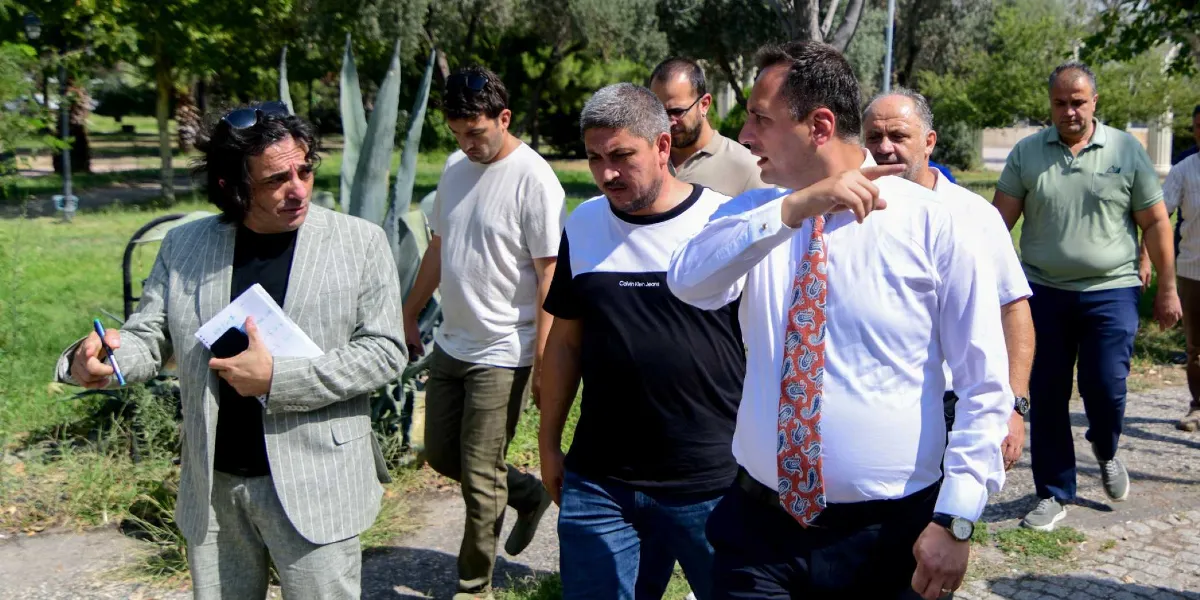 Açık havada, yeşil bir alanda yürüyen bir grup erkek, çevre düzenlemesi veya yeşil alan çalışmaları hakkında incelemelerde bulunuyor. Gruptaki beyaz gömlekli ve desenli kravatlı bir kişi, eliyle ileriyi işaret ederek bir şeyler anlatıyor; yanında ise elinde not defteri ve kalem tutan, çizgili ceketli bir kişi onu dinliyor. Diğer katılımcılar ise onları takip ederek çevreyi gözlemliyor.