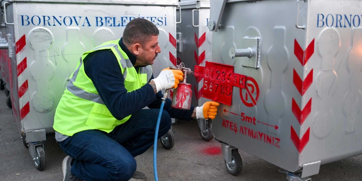 Bornova Belediyesi logolu metal çöp konteynerlerinin yanında diz çökmüş, üzerinde fosforlu sarı iş yeleği ve eldivenleri olan bir belediye çalışanı, elindeki sprey boya tabancasıyla konteyner üzerine numara ve uyarı yazıları yazıyor. Konteynerin üzerinde kırmızı renkli 