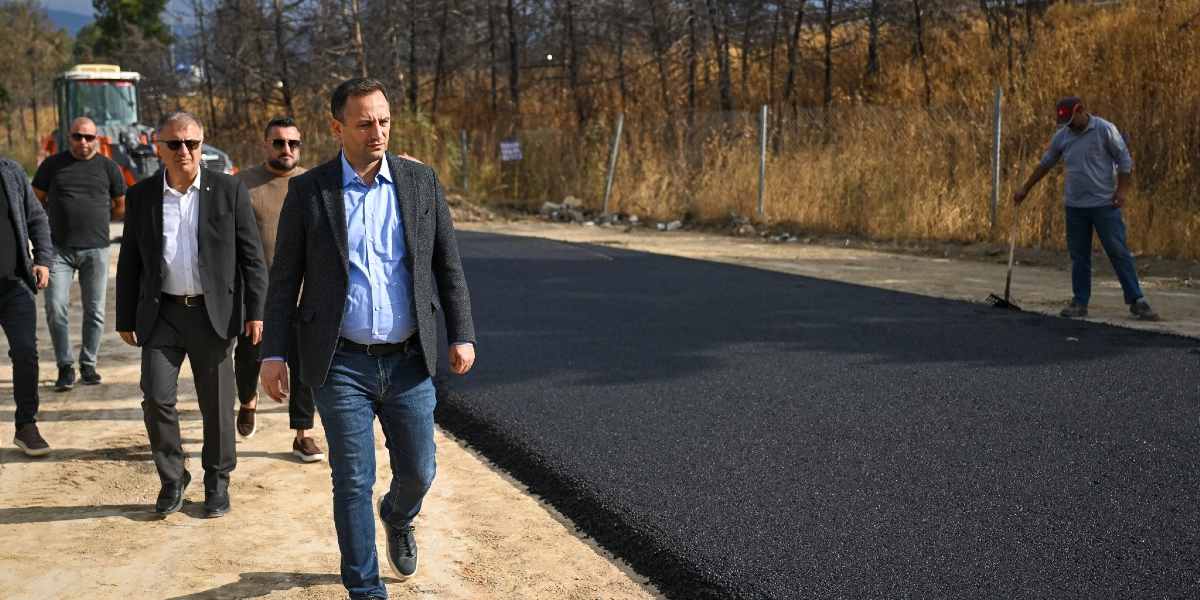 Bornova Belediye Başkanı olduğu düşünülen bir yetkili ve beraberindeki heyet, yeni dökülmüş asfalt yolun kenarında yürüyerek çalışmaları inceliyor. Arka planda kuru otlar ve ağaçlık bir alan bulunurken, sağ tarafta bir işçi elindeki tırmıkla yol kenarındaki düzenleme çalışmalarına devam ediyor.