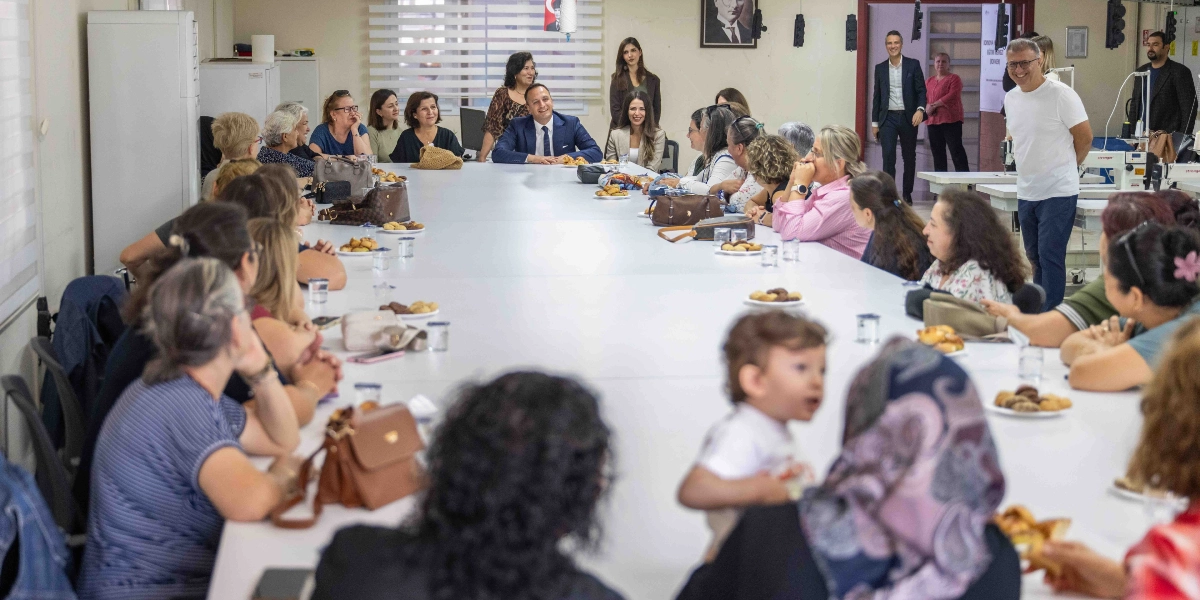 Geniş bir toplantı masasının etrafında toplanmış, çoğunluğu kadınlardan oluşan bir grup insan bir araya gelmiş. Masanın başında takım elbiseli bir erkek ve yanında bir kadın otururken, masanın diğer ucunda beyaz tişörtlü bir erkek ayakta duruyor. Masanın üzerinde tabaklarda kurabiyeler ve bardaklar bulunuyor. Toplantı odasında, masanın etrafında oturanlar arasında küçük bir çocuk da yer alıyor. Arka planda duvarda Atatürk portresi ve Türk bayrağı asılı.