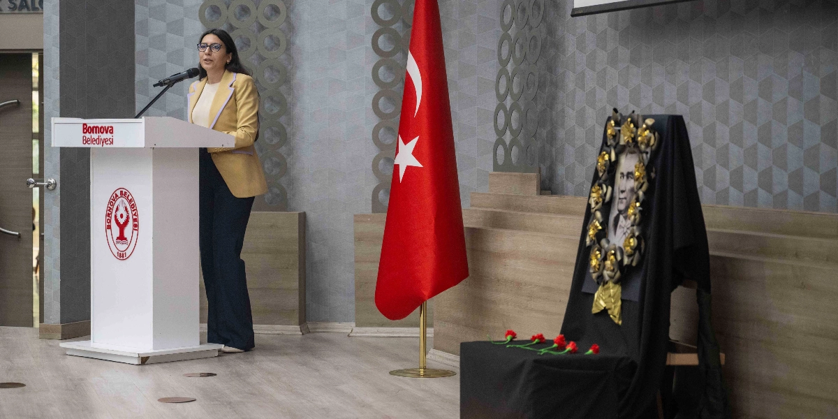 Bornova Belediyesi kürsüsünde konuşma yapan gözlüklü bir kadın, yanında Türk bayrağı ve üzerinde Atatürk portresi ile siyah çelenk bulunan bir anma köşesi yer alıyor.