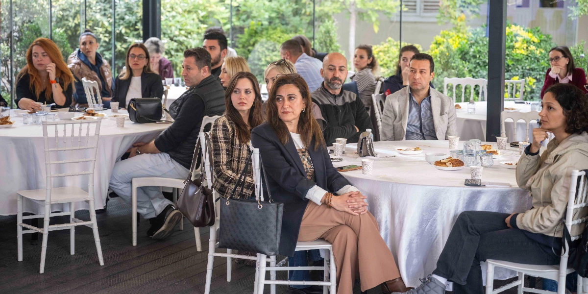 Cam duvarlarla çevrili aydınlık bir salonda, beyaz örtülü yuvarlak masaların etrafında oturmuş, dikkatle dinleyen bir grup insan. Katılımcılar arasında kadın ve erkekler yer alıyor; birçoğu şık ve günlük kıyafetler içinde, masalarda ise içecekler ve atıştırmalıklar bulunuyor. Ön planda, masada oturan ve konuşmacıyı takip eden katılımcılar odak noktasında yer alıyor.