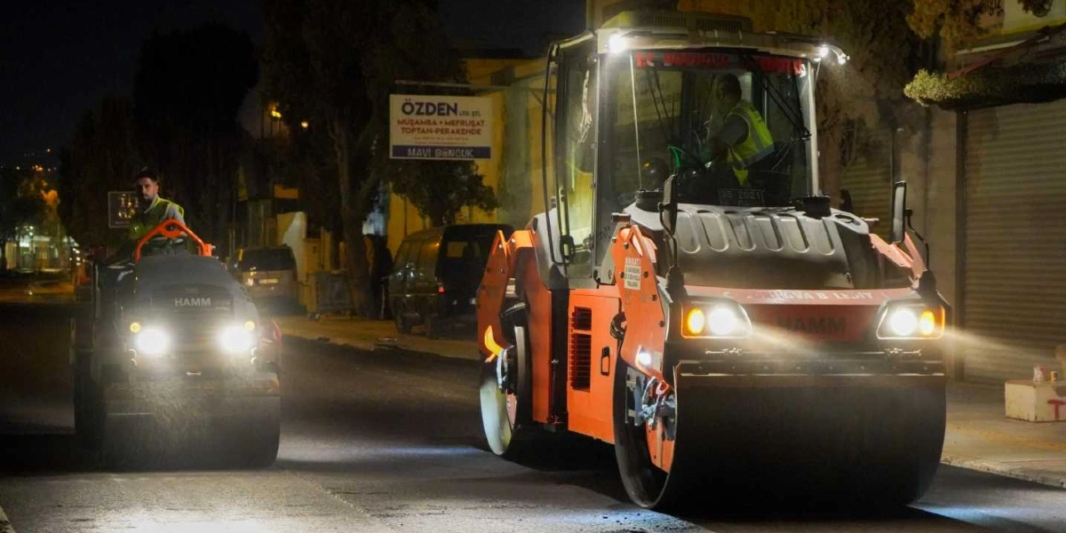 Gece vakti bir cadde üzerinde asfalt çalışması yapan iki adet Hamm marka silindir iş makinesi, üzerlerinde reflektörlü yelek giyen operatörleriyle birlikte ilerliyor. Arka planda dükkan tabelaları ve park halindeki araçlar görülüyor.