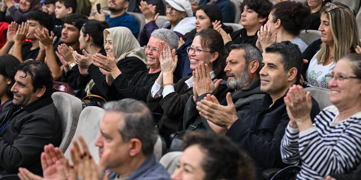 Kapalı bir salonda, farklı yaş gruplarından oluşan kalabalık bir izleyici kitlesi, gülümseyerek ve alkışlayarak bir etkinliği izliyor. Ön planda, çizgili tişörtlü bir kadın, koyu renk ceketli erkekler ve gözlüklü kadınlar dahil olmak üzere çeşitli izleyiciler yer alıyor. Arka sıralarda ise başörtülü kadınlar ve gençlerin de bulunduğu, neşeli bir atmosferin hakim olduğu bir topluluk görülüyor.